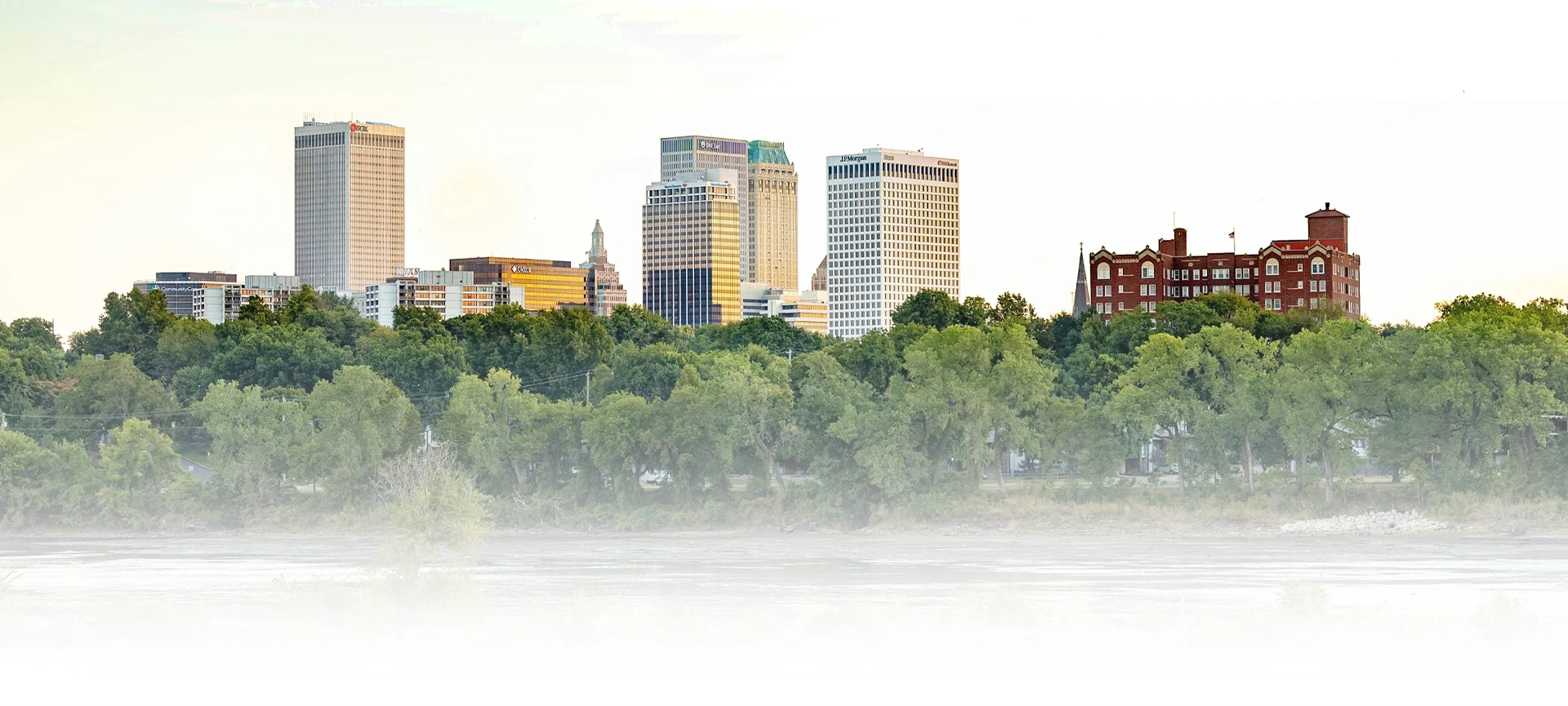 Tulsa city skyline with modern skyscrapers and a historic red-brick building, framed by green trees and a river in the foreground