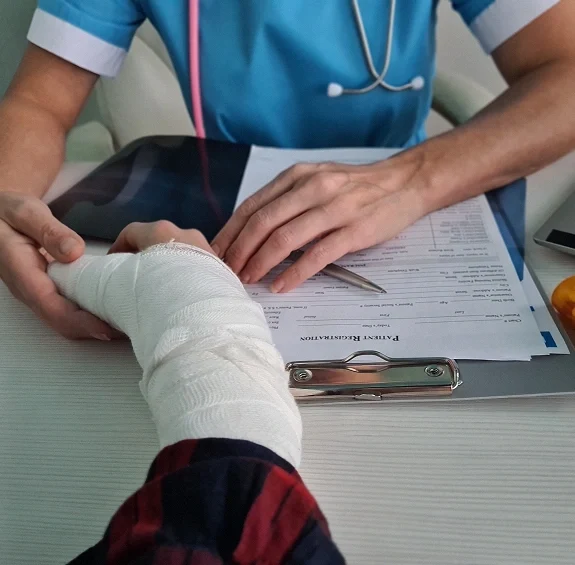Doctor in blue scrubs holding a patient's heavily bandaged hand while reviewing paperwork on a clipboard