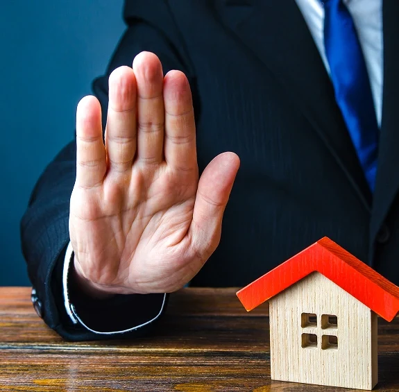 Person in suit holding hand up in a stop gesture next to a small wooden house model on a wooden surface