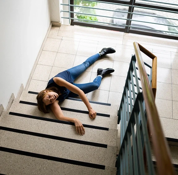 Woman lying on stair landing appearing injured or unwell, holding onto the step railing indoors near a window