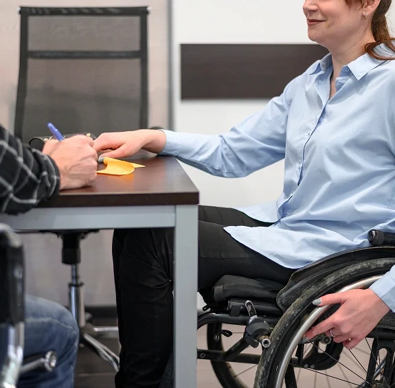 A woman in a wheelchair is engaged in a conversation with a person sitting across a desk, writing on a paper
